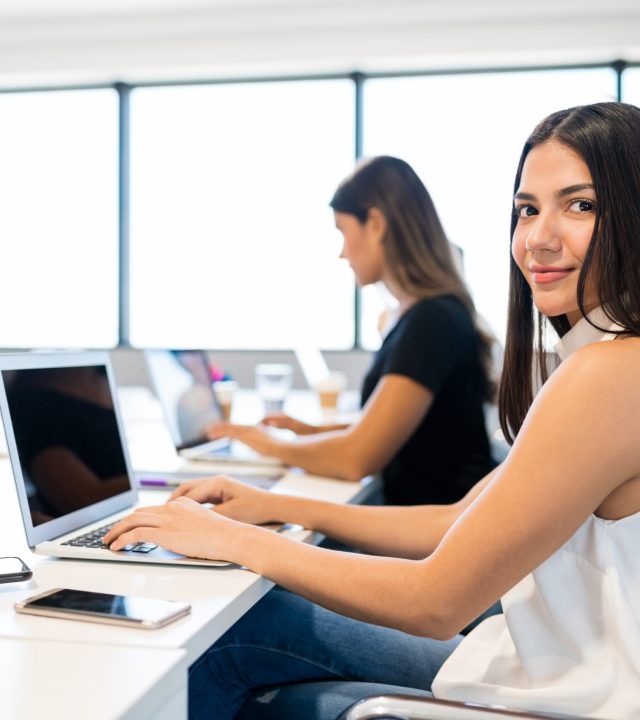 Smiling businesswoman working on laptop while sitting with colleagues at desk in office